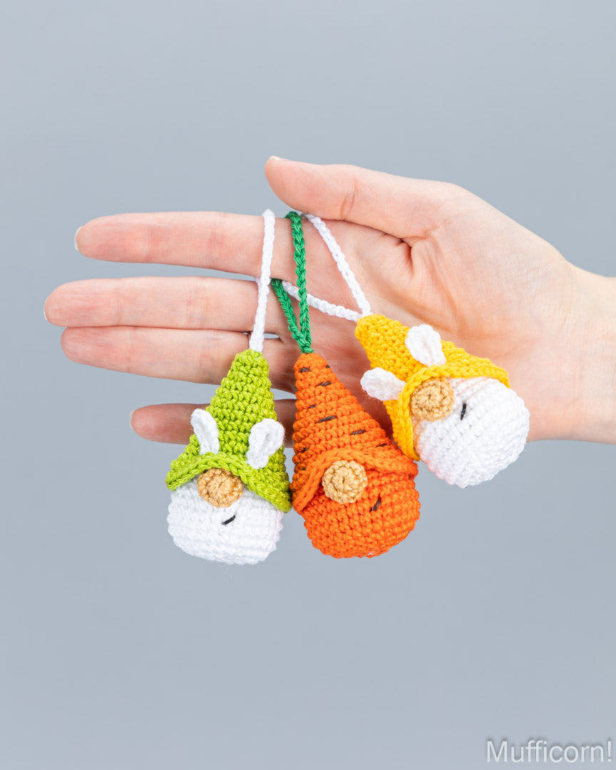 Three crocheted carrot-shaped ornaments with white bunny faces are held in a person's hand against a plain light gray background. The ornaments have green, orange, and yellow tops, with white bunny ears and snouts, and are attached to white and green hanging loops.