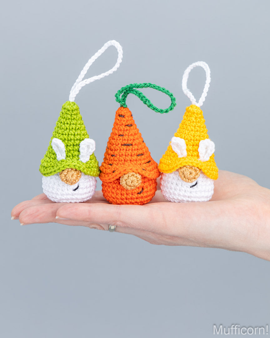 Three small crocheted Easter gnome ornaments are held in a person's hand against a plain light gray background. The left gnome wears a green hat, the center gnome wears an orange carrot-shaped hat, and the right gnome wears a yellow hat.