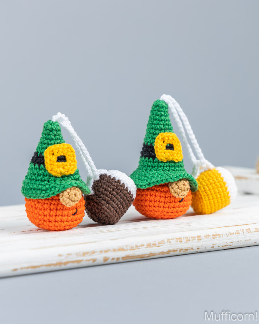 Two crocheted gnomes with green hats, orange bodies, and white hanging loops, styled on a white wooden surface against a soft gray background.