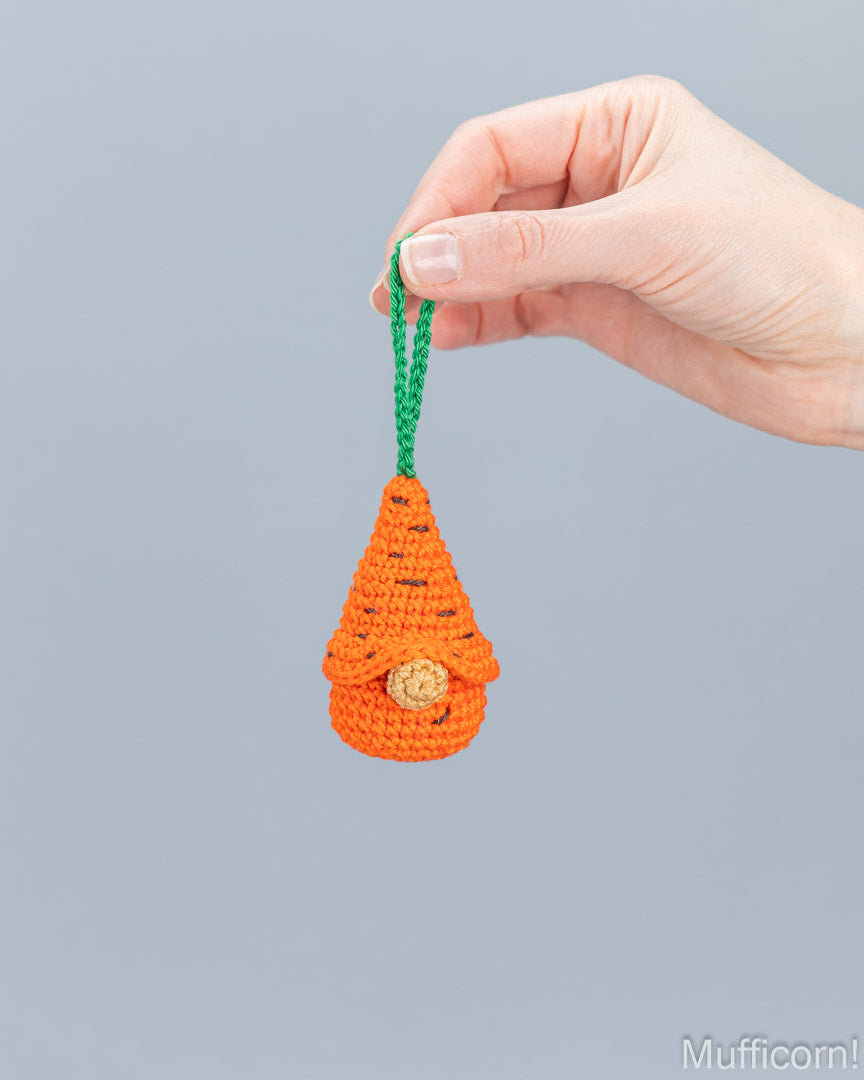 A hand holding a small crocheted carrot-shaped ornament with a green hanging loop and a wooden button nose, against a plain light gray background.