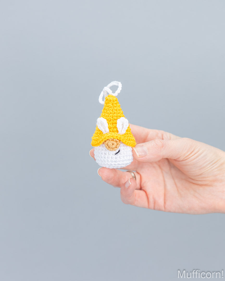 A hand holding a small crocheted bunny gnome keychain against a solid gray background.