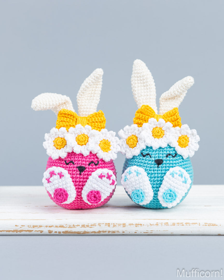 Two crocheted Easter bunny eggs side by side on a white wooden surface against a light gray background. The left egg is pink with white daisy accents and a yellow bow, and the right egg is blue with white daisy accents and a yellow bow.