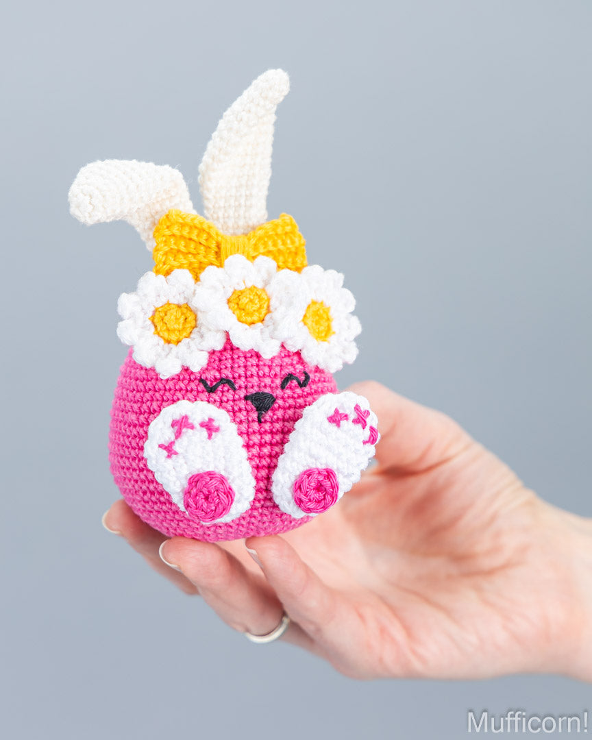 A crocheted pink bunny egg ornament with white daisy flowers, white ears, and a yellow bow, held in a person's hand against a plain gray background.