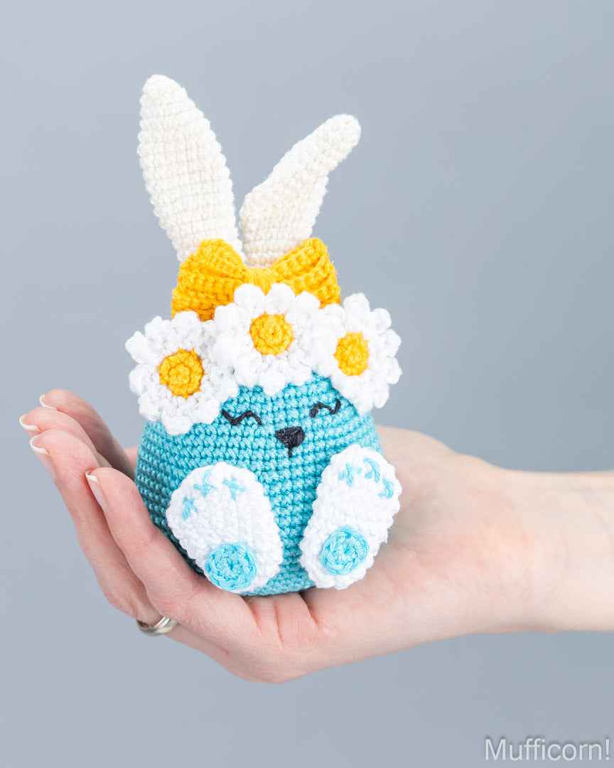 A crocheted bunny doll with white ears, a yellow bow, white daisy flowers, and white feet, held in a person's hand against a plain gray background.