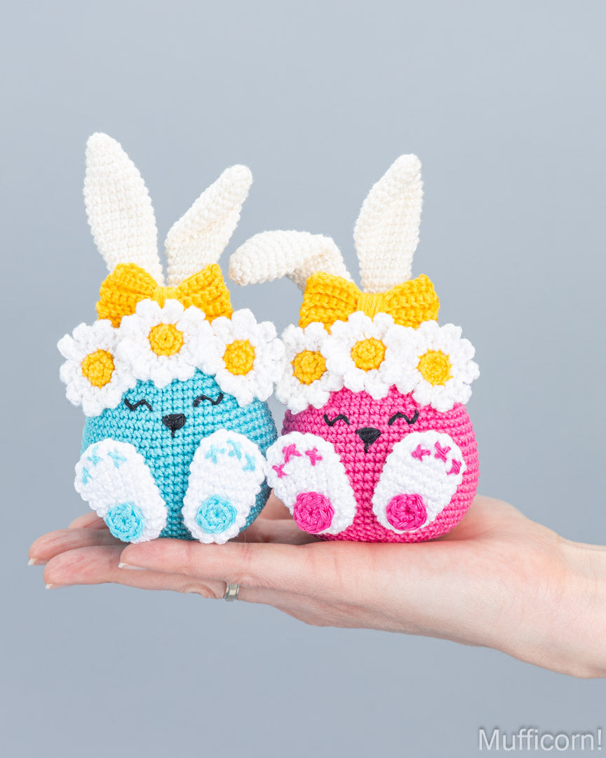 Two crocheted bunny amigurumi dolls are held in a person's hand against a plain gray background. One bunny is light blue with white daisy flowers and a yellow bow, and the other is pink with white daisy flowers and a yellow bow.