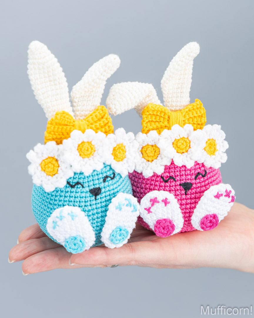 Two crocheted Easter bunny amigurumi dolls in light blue and pink, each with white daisy flower crowns, yellow bows, and white bunny ears, displayed on a person's hand against a solid gray background.