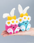 Two crocheted bunny characters in pink and blue, each with white daisy-like flower crowns, yellow bows, and white bunny ears, displayed on a person's hand against a plain gray background.