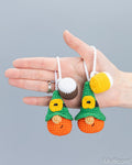 Two small crocheted gnome ornaments are held in a person's hand against a plain light gray background. Each ornament has an orange body, a green hat, yellow facial details, and white hanging loops.