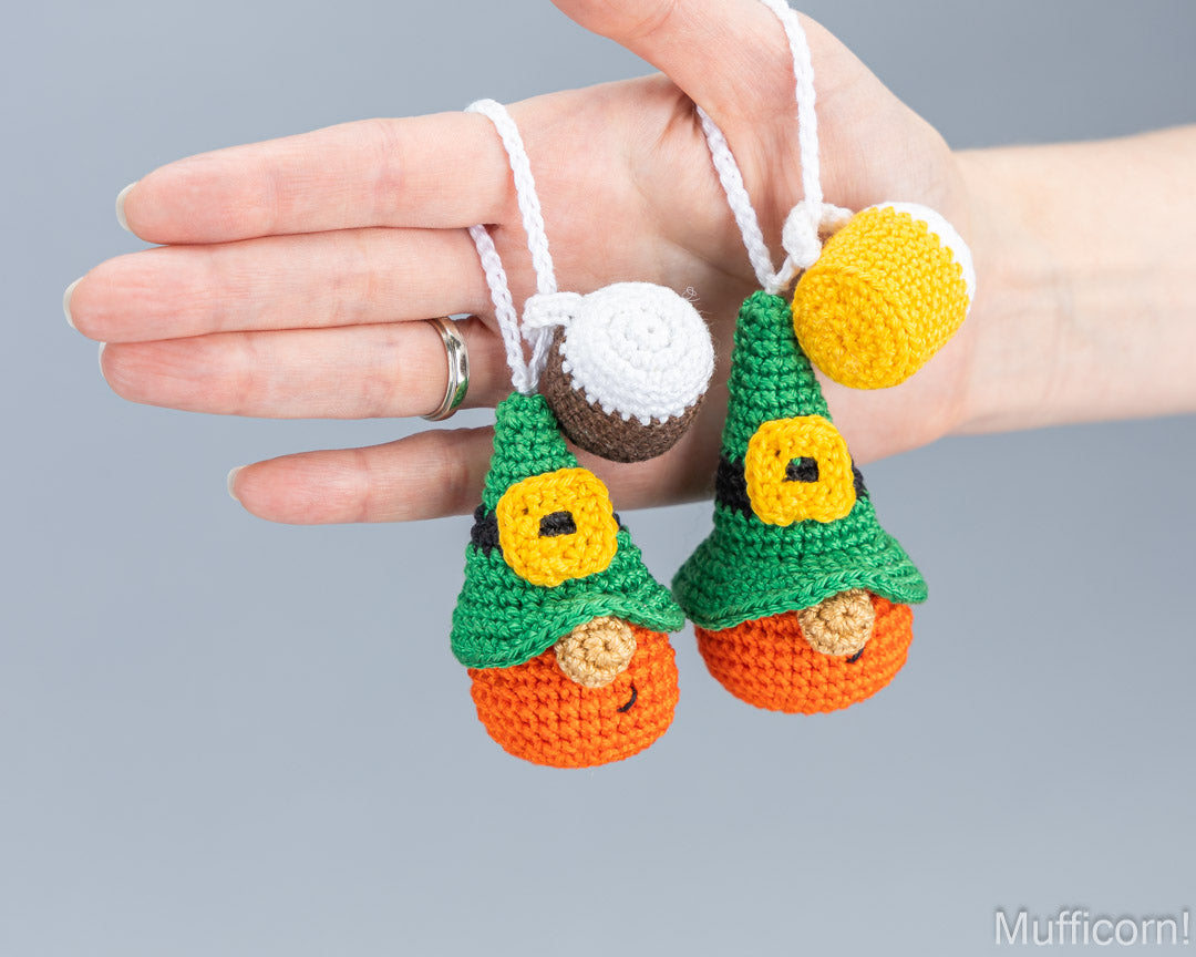 A close-up of a hand holding two crocheted leprechaun ornaments against a plain gray background. The ornaments have green pointed hats, orange bodies, yellow faces, and white hanging loops.
