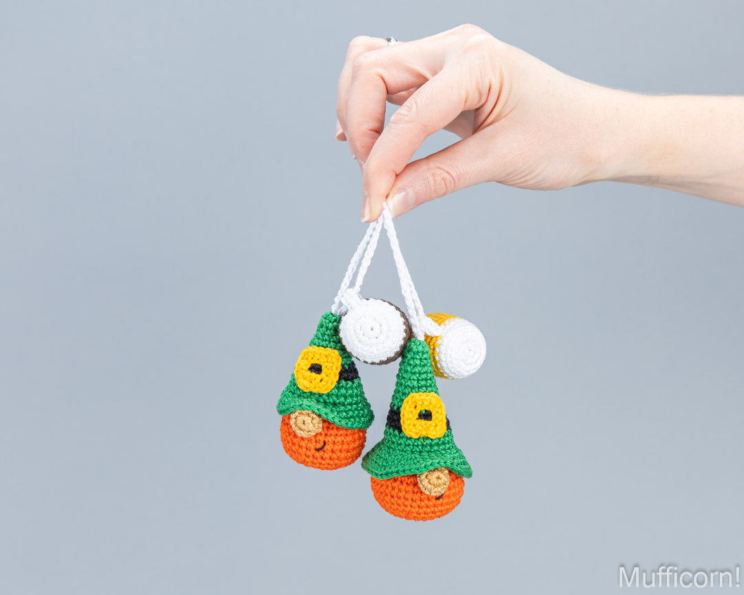 A hand holds two small crocheted ornament shapes shaped like leprechauns, featuring green hats, yellow buttons, orange bodies, and white hanging loops, against a plain light gray background.