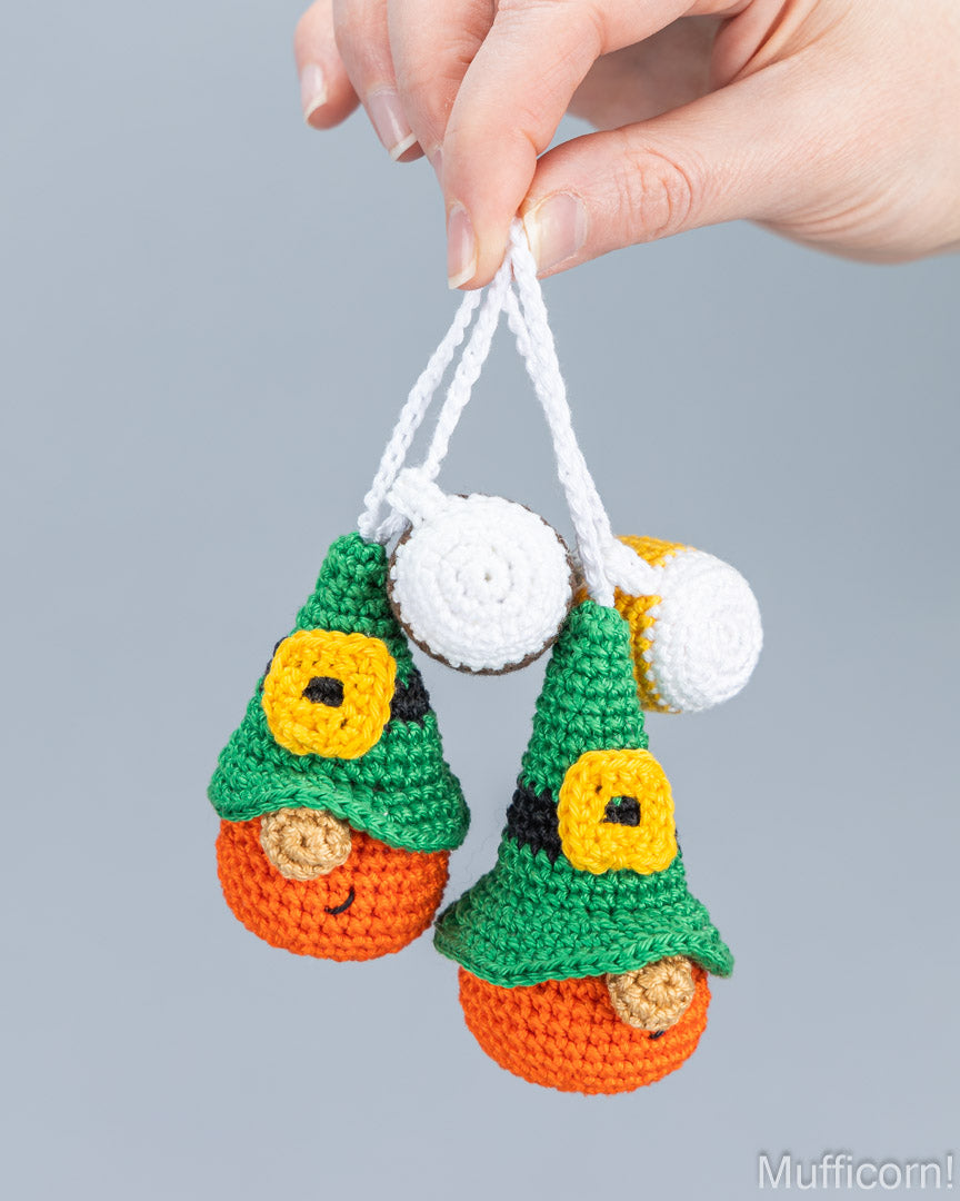 A hand holds two small crocheted gnome ornaments against a plain light gray background. The ornaments have green pointed hats, yellow square faces, orange bodies, and white hanging loops with white pom-poms.