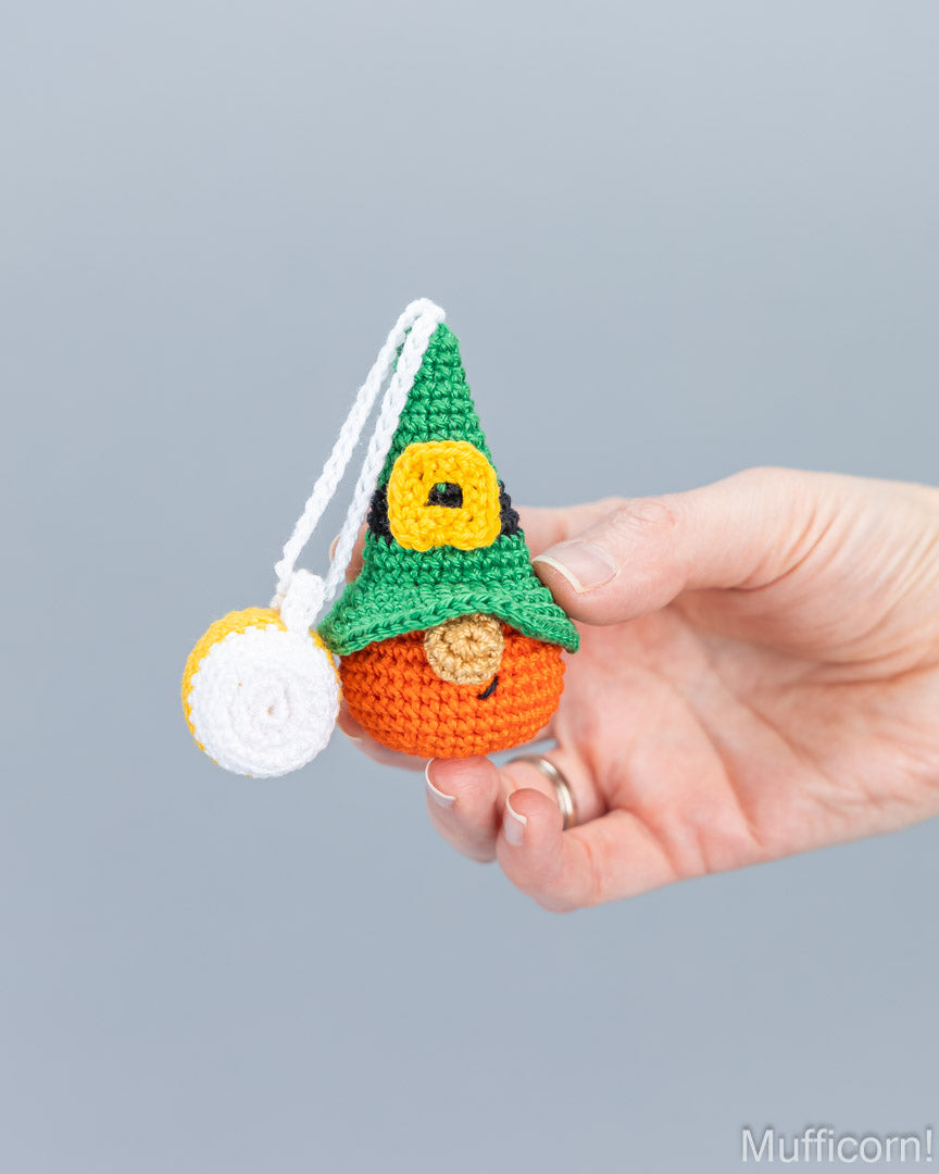 A hand holds a small crocheted gnome ornament against a plain gray background. The gnome has a green pointed hat with a yellow band, an orange body, a small brown nose, and a white hanging loop with a white ball accessory.