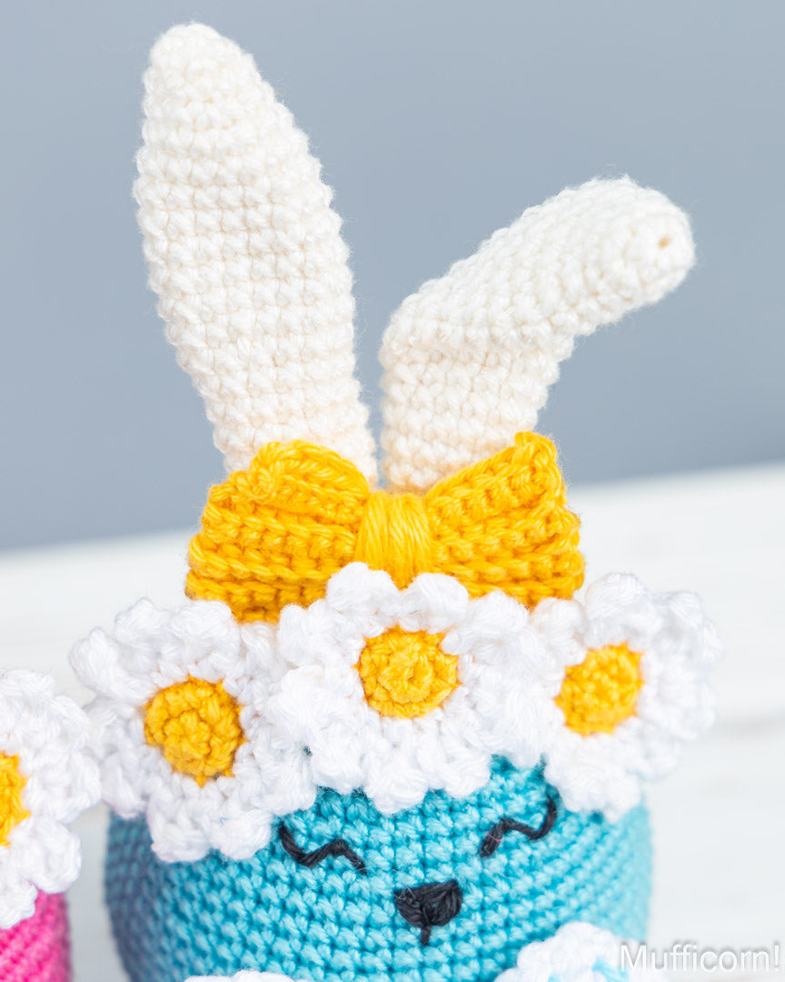 A close-up of a crocheted bunny character with long white ears, a yellow bow, white daisy flowers with yellow centers, and a turquoise body against a soft gray background.