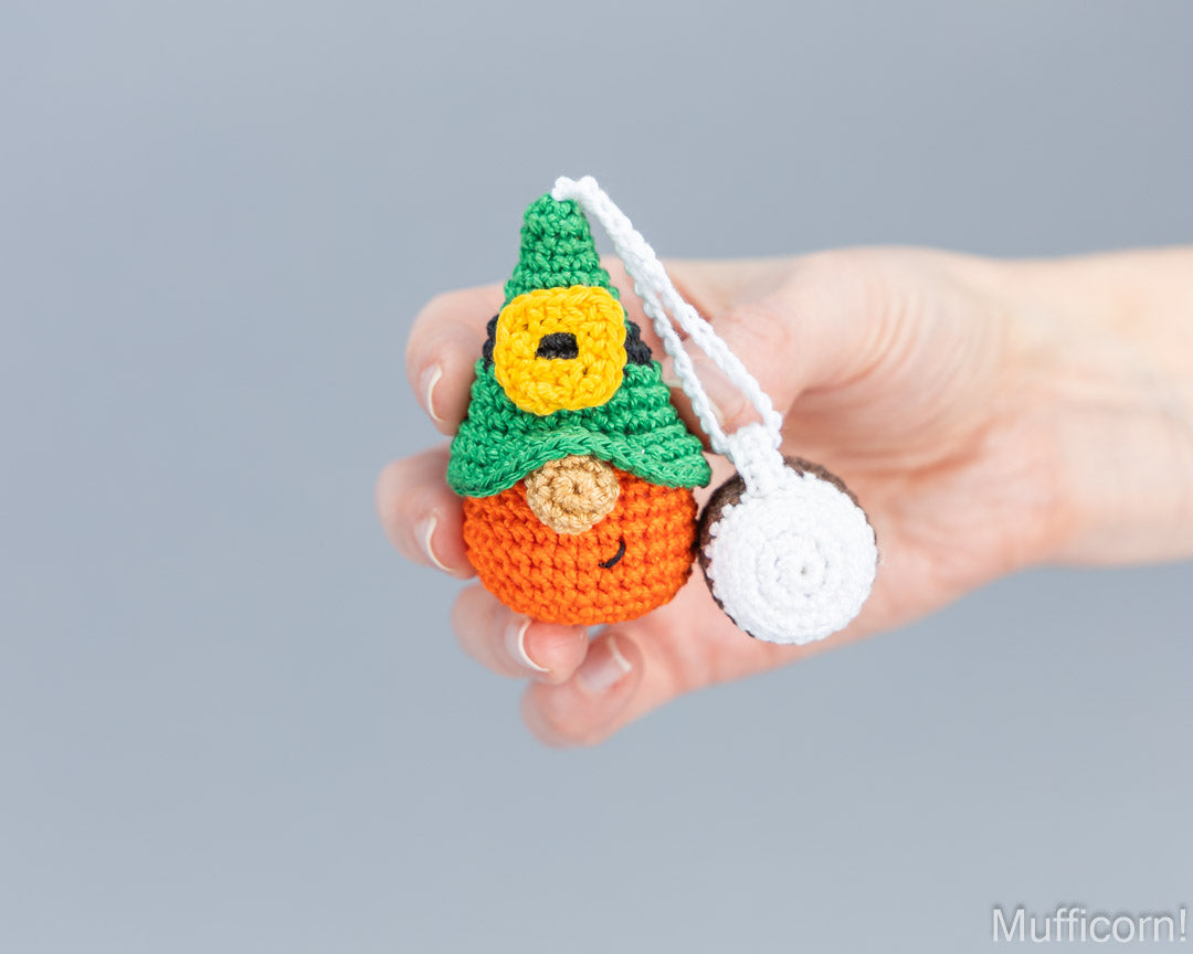 A hand holding a small crocheted gnome keychain with a white circular keyring against a solid gray background.