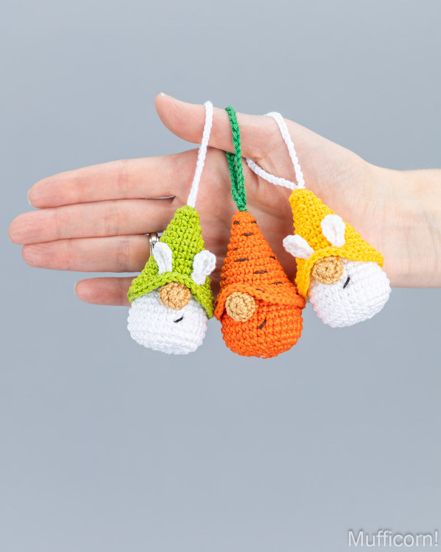 Three small crocheted bunny-shaped keychains or charms are held in a person's hand against a plain light gray background. Each bunny has a different colored hat and body: green, orange, and yellow, with white faces and ears.
