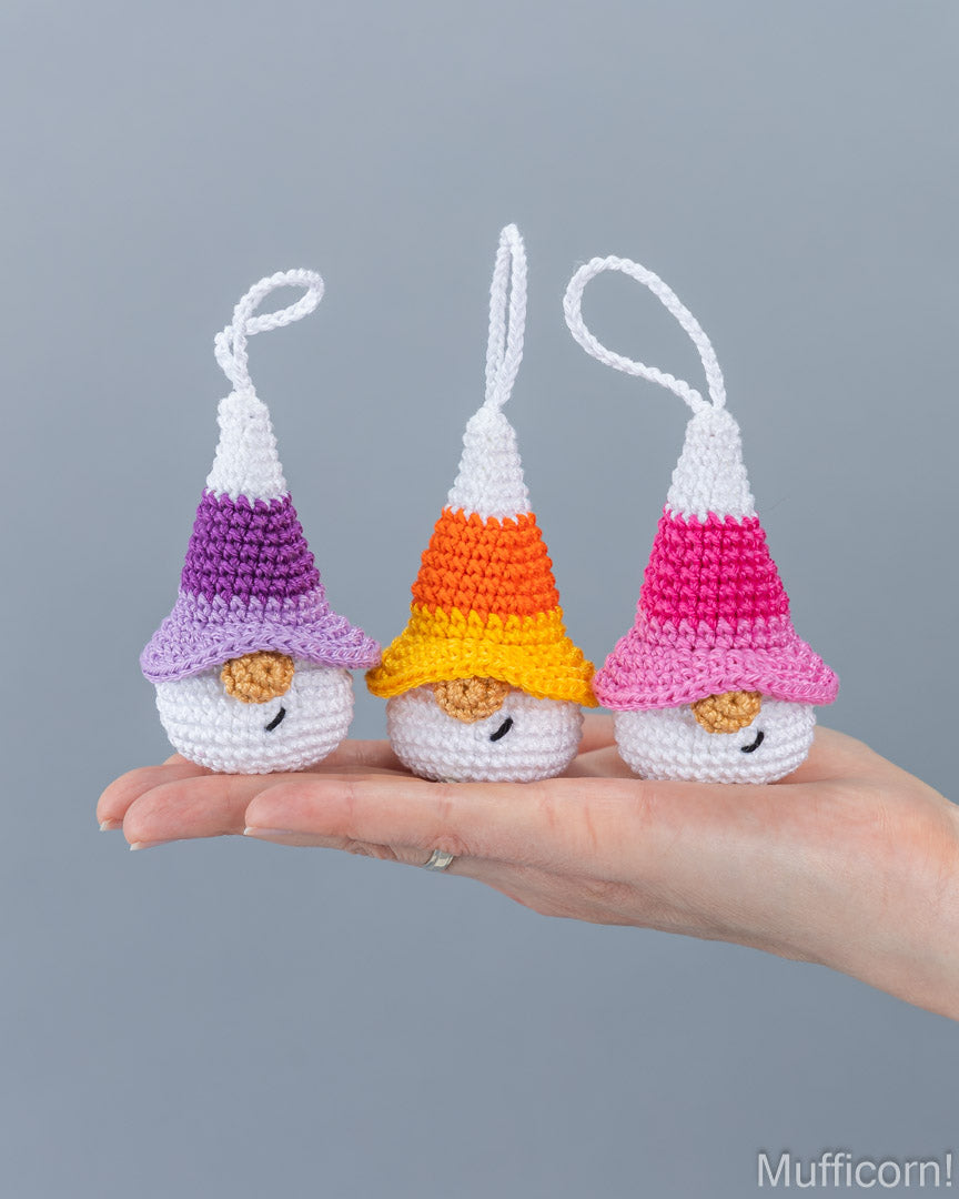 Three small crocheted gnomes with hanging loops are displayed on a person's hand against a plain gray background. Each gnome has a striped hat in different color combinations: purple and white, orange and yellow, and pink and white.