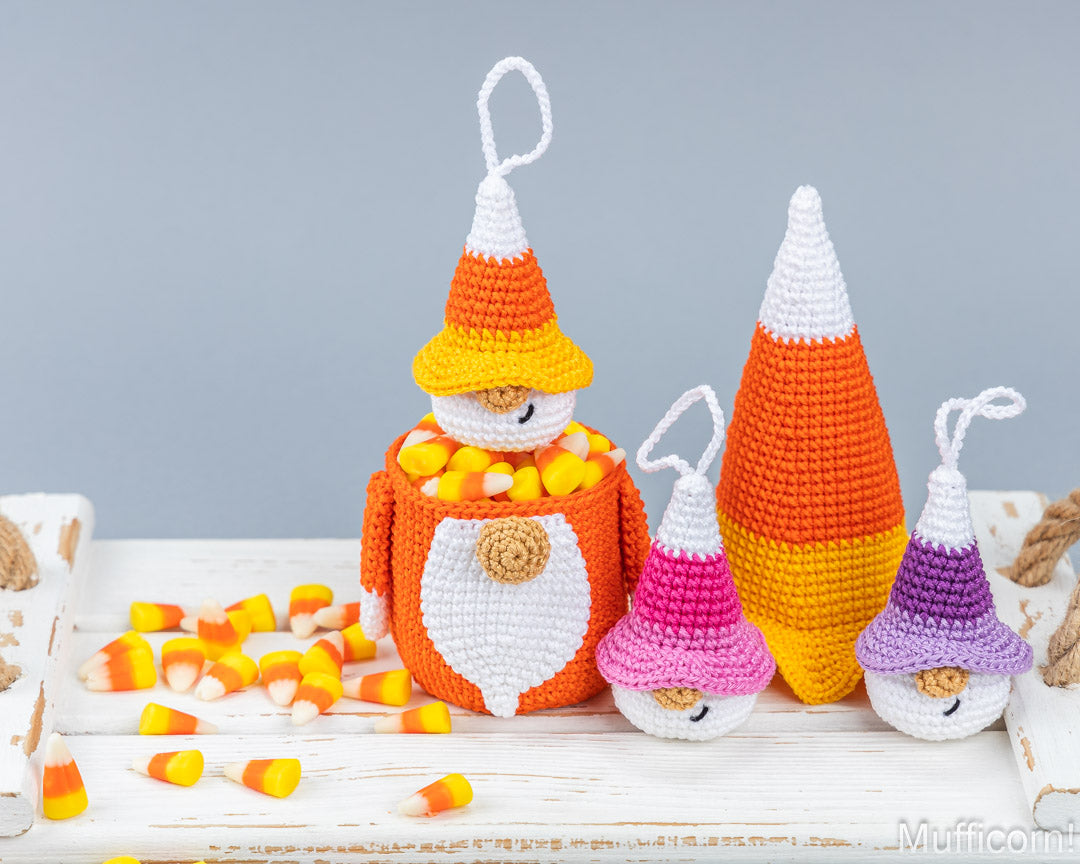 Crocheted amigurumi Halloween decorations including a large candy corn pot filled with candy corn, a tall candy corn tree, and two small candy corn figures, arranged on a white wooden surface with scattered candy corn against a gray background.