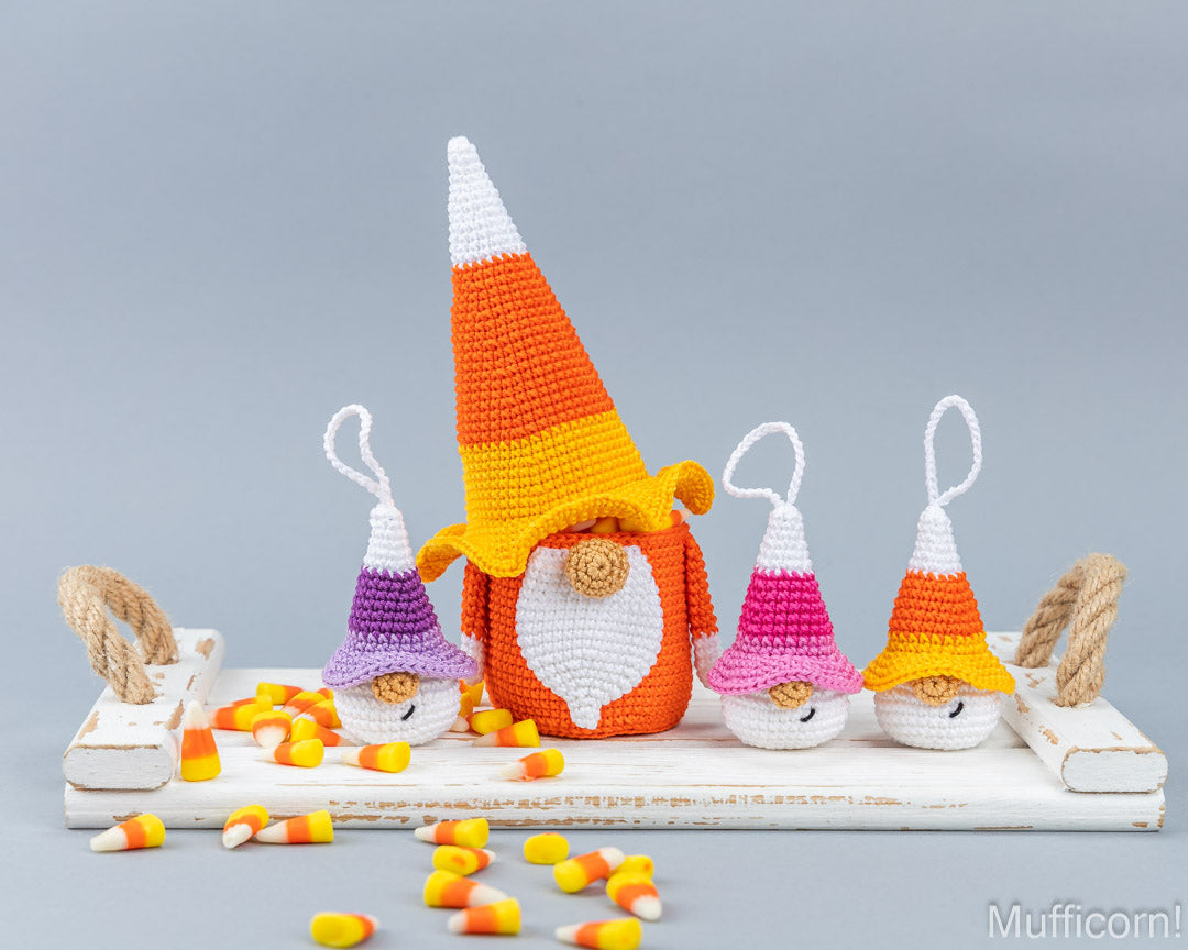A crocheted Halloween ornament set featuring a large central candy corn gnome and three smaller matching gnomes on a white wooden platform, with scattered candy corn pieces on a gray studio background. The crocheted hats are striped in orange, yellow, white, purple, pink, and orange-yellow.