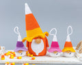 A crocheted gnomes set styled on a white wooden surface with candy corn. The set includes one large gnomedom in the center wearing a classic orange, yellow, and white corn cob hat, flanked by four smaller gnomodoms wearing purple, pink, and orange/yellow striped hats.