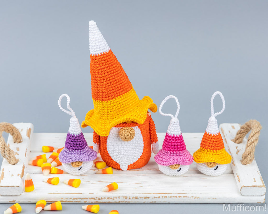 Three crocheted candy corn gnomes with hanging loops, displayed on a white rustic wooden tray with scattered candy corn pieces against a solid gray background.
