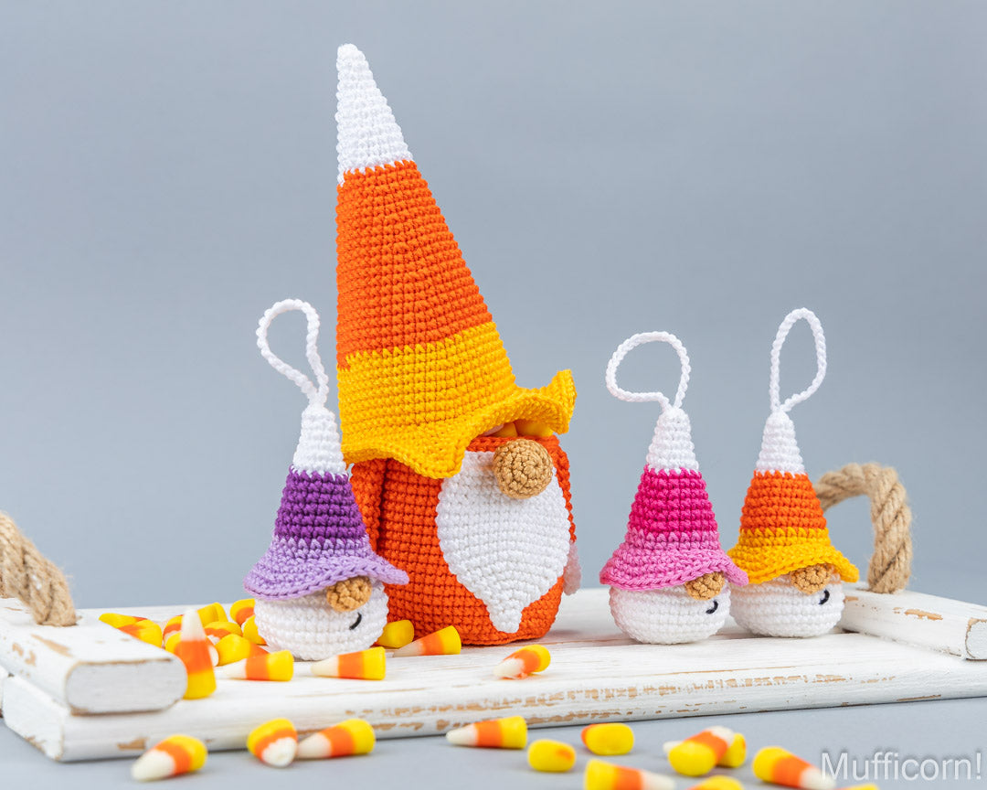 Three crocheted candy corn gnomes with hanging loops, arranged on a white wooden tray with scattered candy corn against a soft gray background.