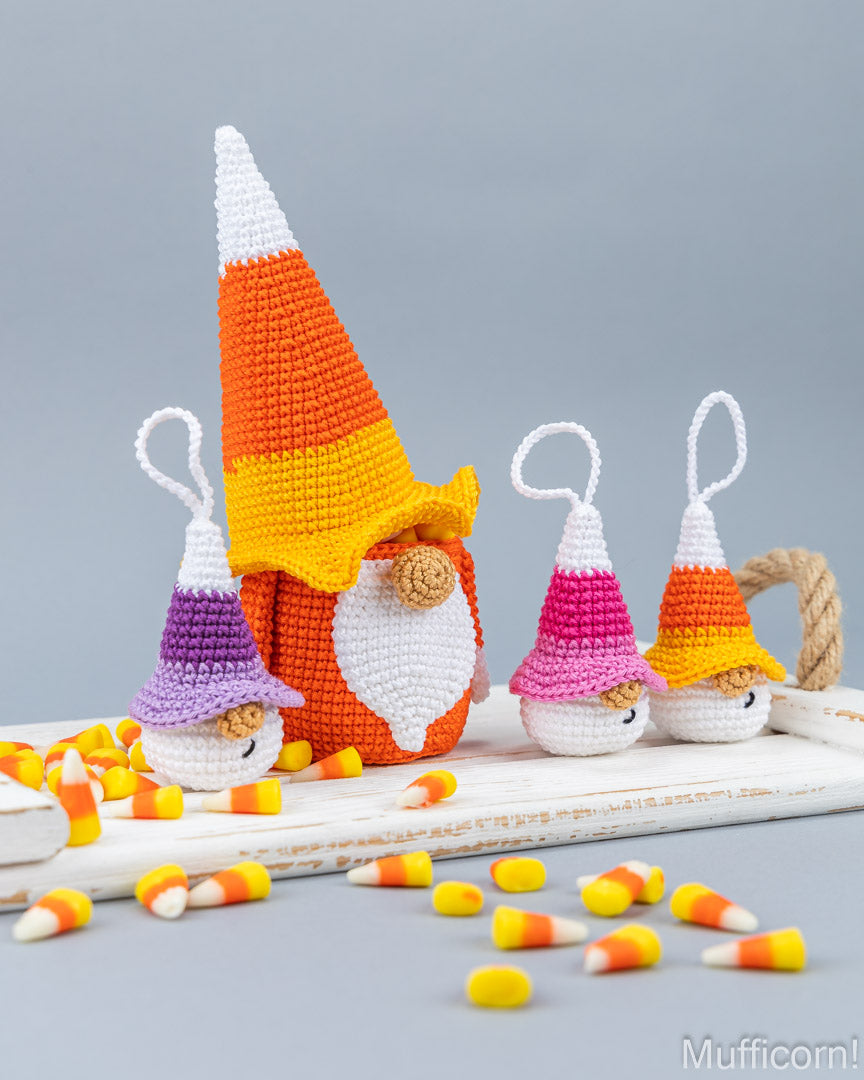 Three crocheted Halloween gnomes styled like candy corn, displayed on a white wooden board with scattered candy corn pieces on a gray background. The central gnome is larger and wears a purple, white, and orange hat, while the two smaller gnomes on either side wear pink, white, and orange hats.