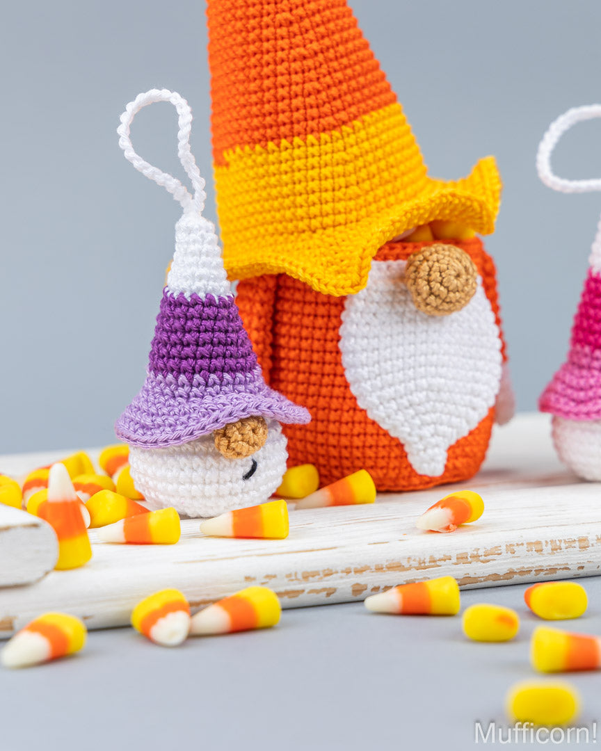 Handmade crocheted Halloween gnomes styled with candy corn on a white wooden surface against a gray background. The main gnome wears an orange and yellow pointed hat and a white beard, while a smaller gnome wears a purple hat.