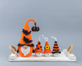 Four crocheted Halloween gnomes in orange, black, and white are arranged on a white wooden tray with rope handles against a smooth gray studio background. A large gnome stands on the left, while three smaller gnomes sit to the right.