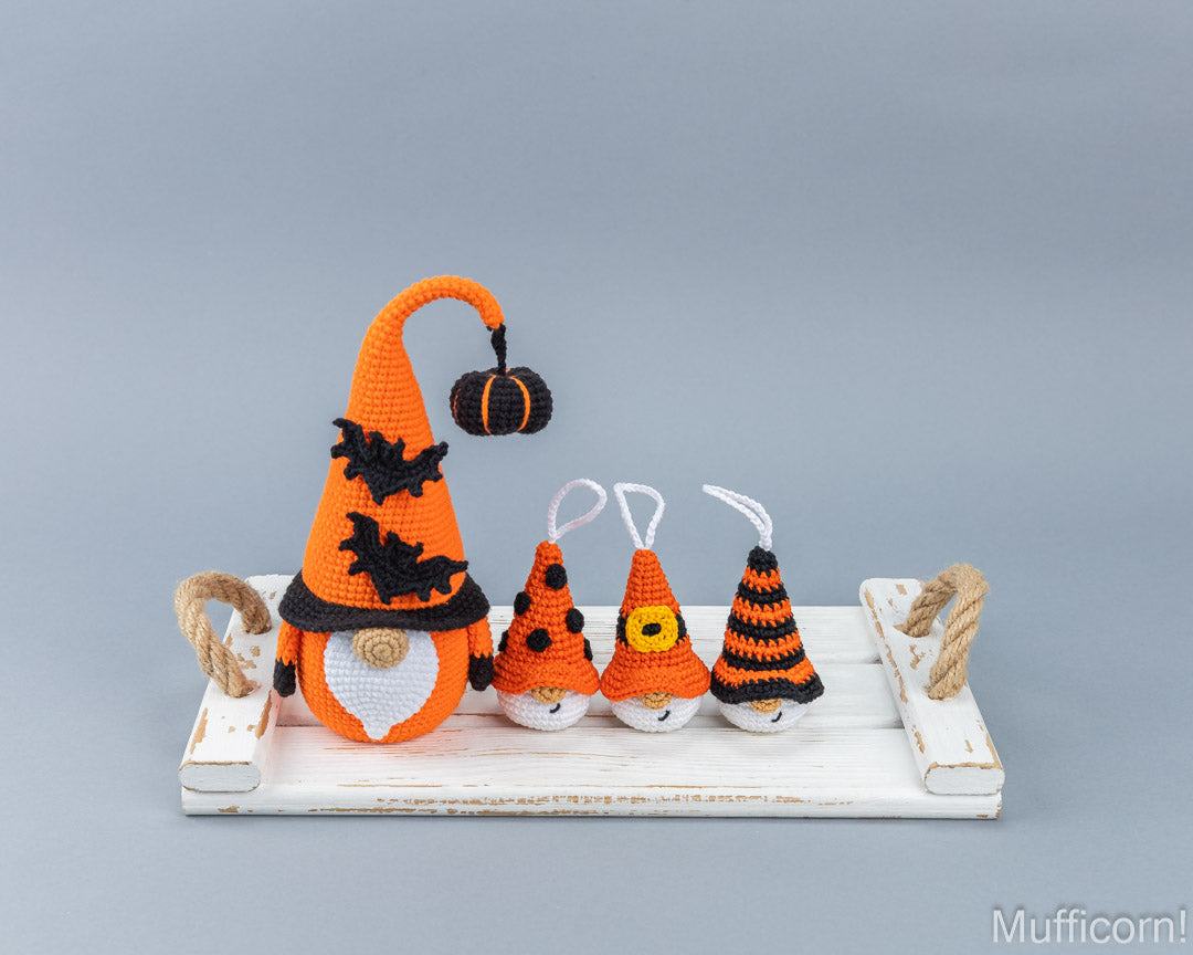A set of crocheted Halloween gnome ornaments displayed on a distressed white wooden tray against a solid gray background. The set includes one large orange gnome with a tall pointed hat and a small hanging pumpkin, and four smaller orange and black striped gnomes.