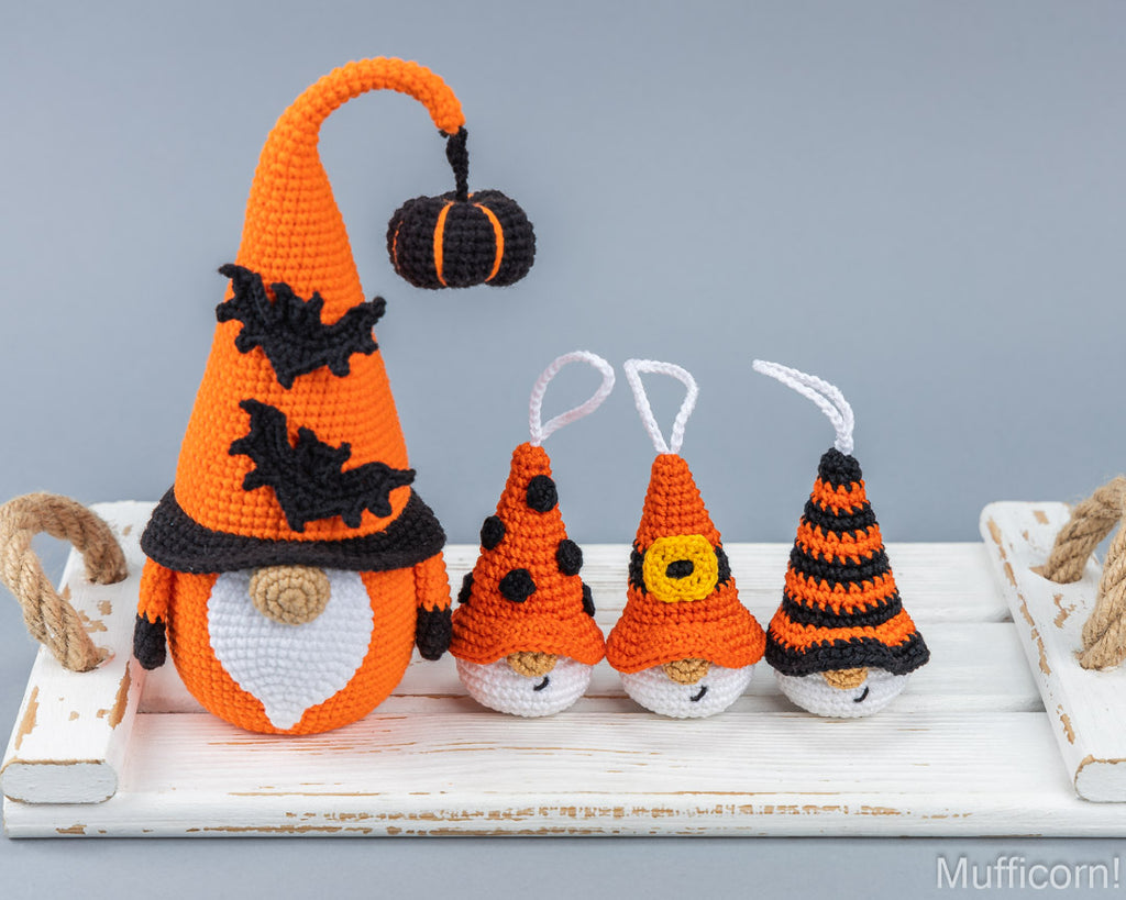 Four crocheted Halloween gnomes in orange, black, and white are displayed on a white wooden tray against a solid gray background. The largest gnome on the left wears a tall orange hat with black bats and a small hanging pumpkin, while the three smaller gnomes on the right wear pointed orange and black hats.