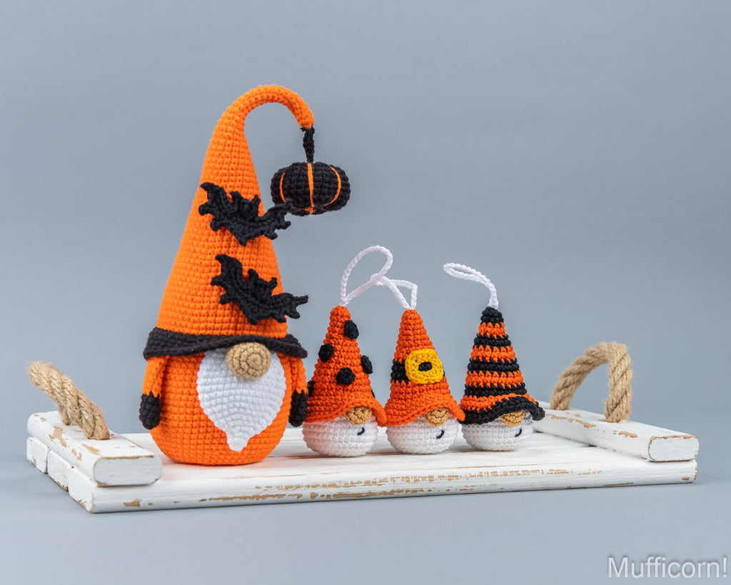 Four crocheted Halloween gnomes in orange, black, and white, arranged on a distressed white wooden tray with rope handles against a solid gray studio background.