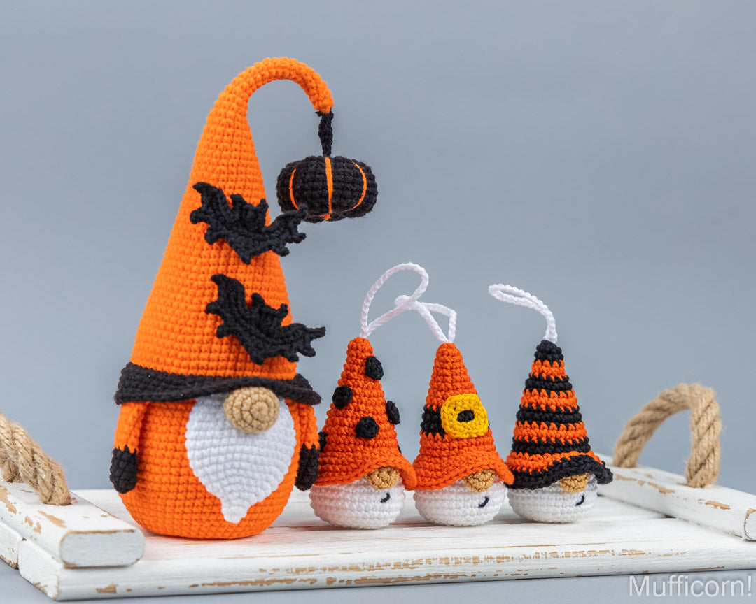 Four crocheted Halloween gnomes in orange and black, displayed on a white rustic wooden tray against a solid gray background.