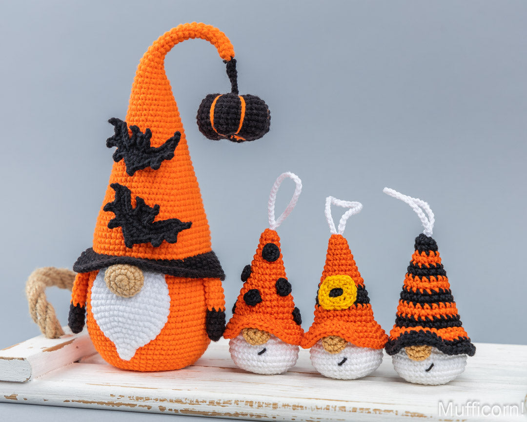 Four crocheted Halloween gnomes in orange, black, and white, including one large gnome with a long hat and a small pumpkin on the tip, and three smaller gnomes with striped and polka-dot hats, displayed on a white wooden surface against a gray background.