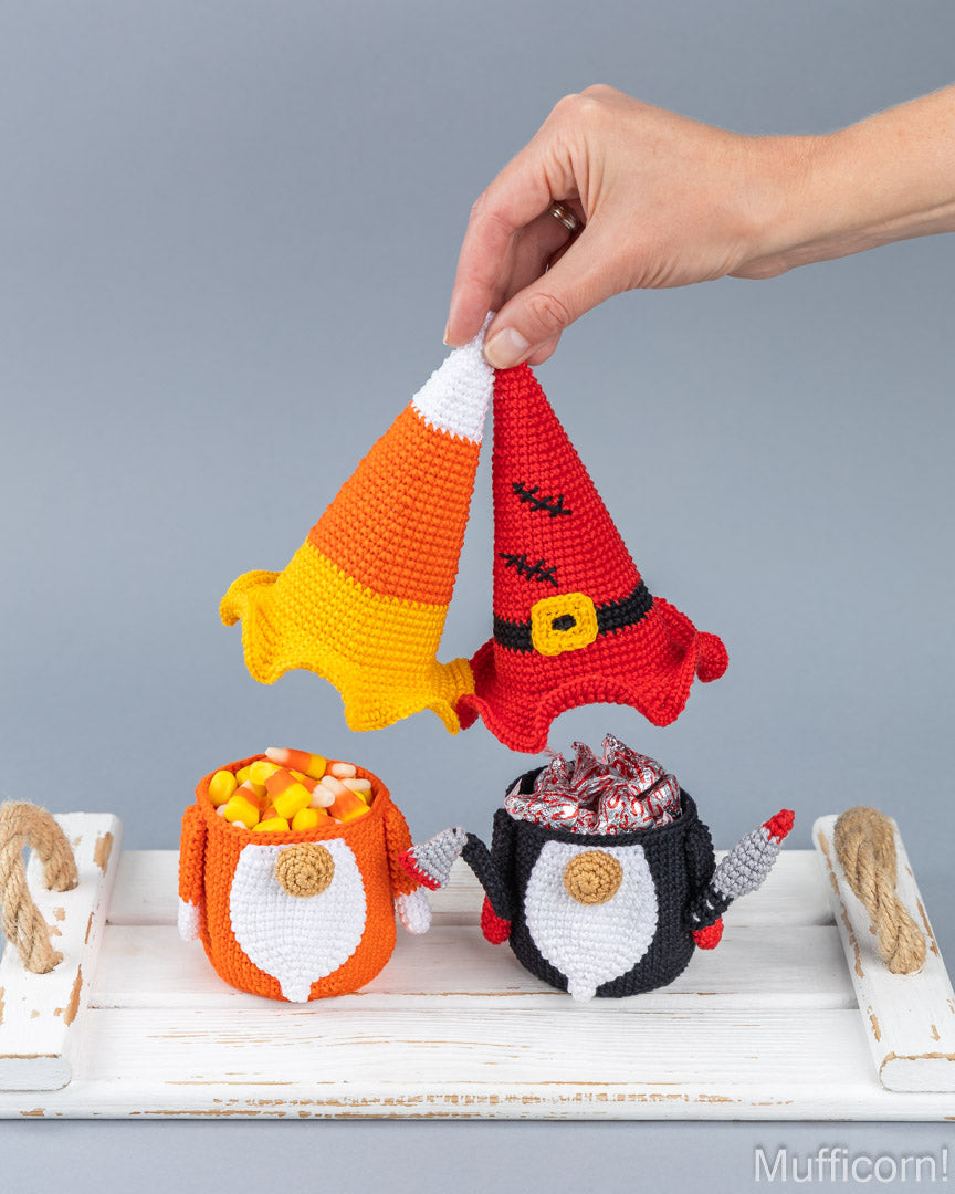 A hand holds two crocheted gnome hats above three small crocheted gnome pots on a white wooden surface against a gray background. The hats are orange-and-white candy corn and red with black and gold trim.