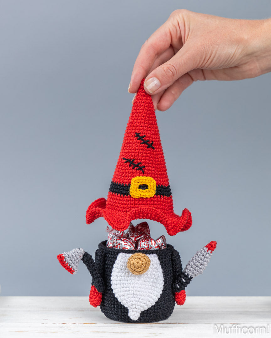 A crocheted gnome figurine with a tall red hat, black and white body, and small red-and-white arms, holding wrapped candies in its mouth. A human hand is lifting the hat, and the product is photographed against a plain light gray background on a white surface.