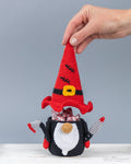 A crocheted gnome figurine with a tall red hat, black and white body, and small red-and-white arms, holding wrapped candies in its mouth. A human hand is lifting the hat, and the product is photographed against a plain light gray background on a white surface.