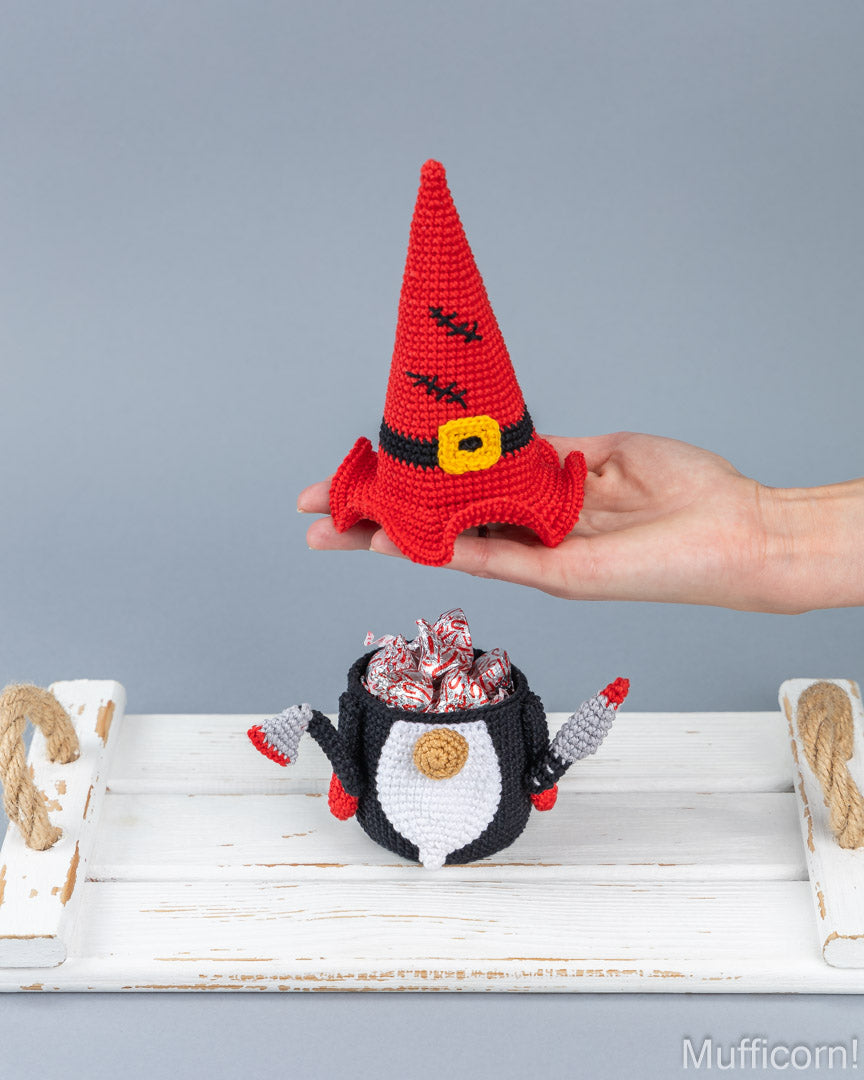 A crocheted gnome-shaped candy pot filled with wrapped candies sits on a white wooden surface, while a hand holds a red gnome hat above it against a gray studio background.