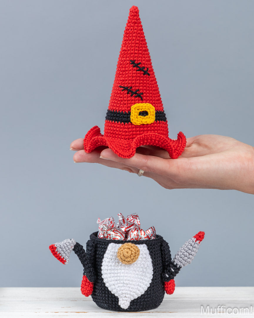 A crocheted red witch hat held in a person's hand above a crocheted halloween gnome candy keeper filled with wrapped candies, against a plain light gray background.