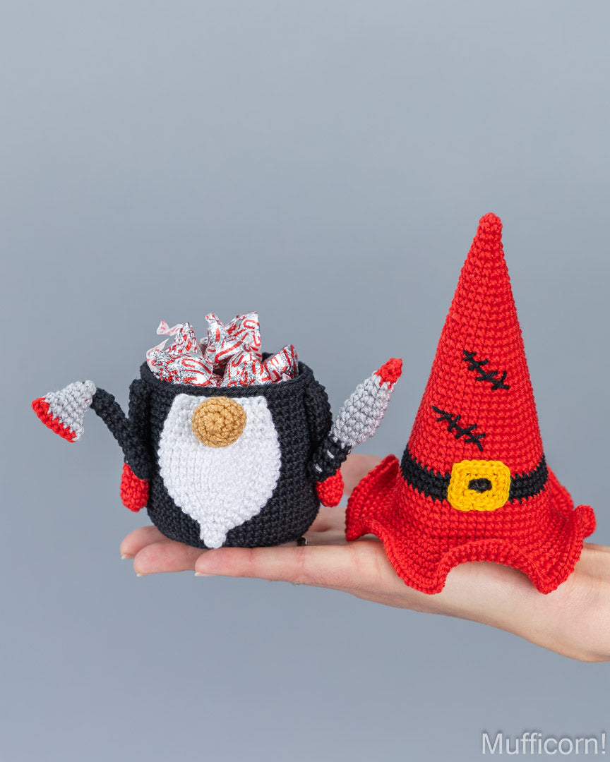 A hand holds two crocheted halloween-themed items against a plain gray background: a black-and-white gnome-shaped candy holder filled with red-and-white wrapped candies, and a red witch hat with black details and a yellow belt buckle.