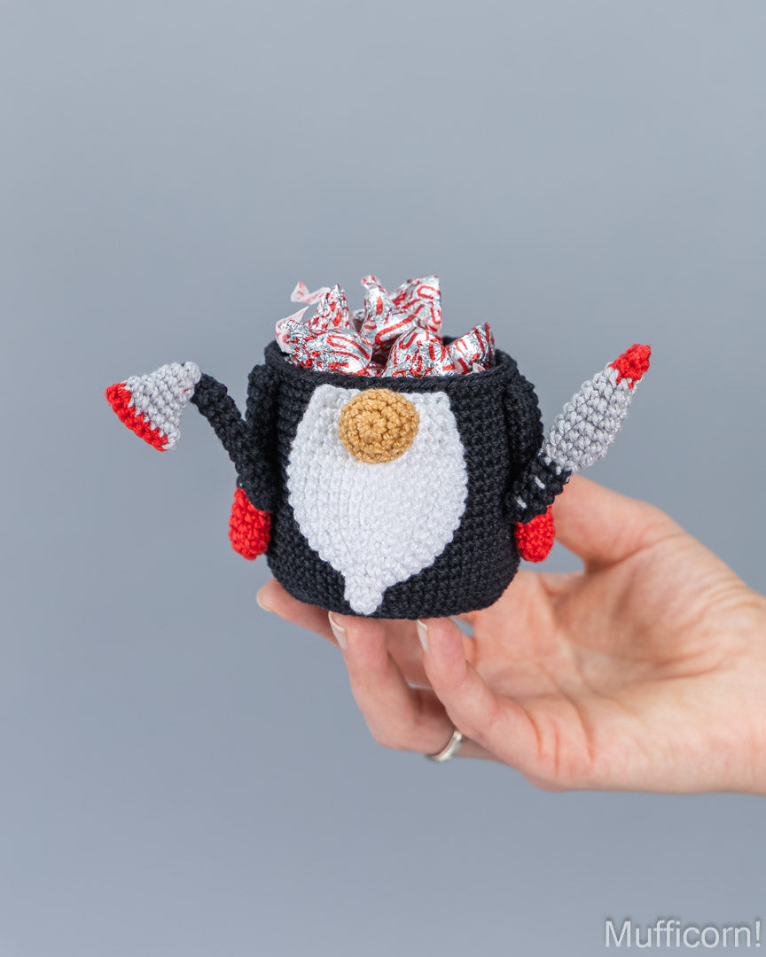 A hand holding a crocheted gnome-shaped candy holder filled with wrapped candies, against a plain gray background.