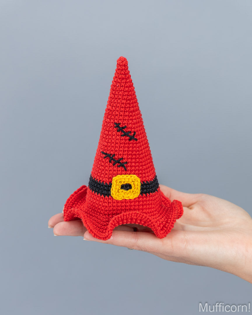 A small crocheted red witch hat with black stitched details and a yellow buckle accent, held in a person's hand against a plain gray background.