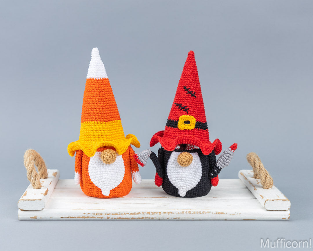 Two crocheted Halloween gnomes standing on a white wooden platform against a gray background. The gnome on the left wears a candy corn hat in orange, yellow, and white with a white beard.