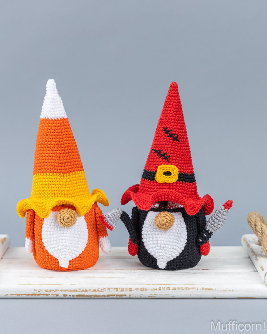 Two crocheted Halloween gnomes displayed side by side on a white wooden surface against a solid gray background. The gnome on the left is styled like a candy corn with orange, yellow, and white sections, and a white beard.