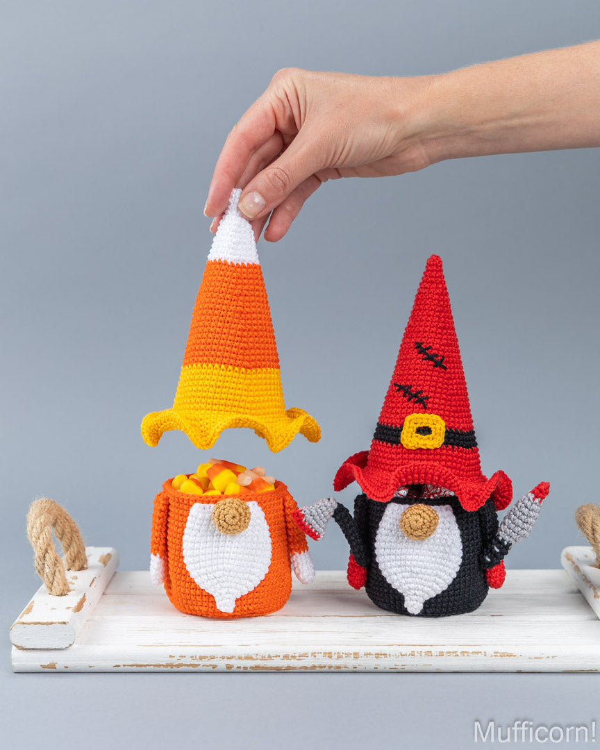Two crocheted Halloween gnomes on a white wooden tray against a gray background. One gnome wears a candy corn hat and orange pot filled with small orange and yellow candies.