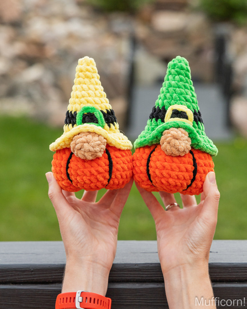 Two crocheted gnome pumpkins held up by two hands outdoors. One pumpkin has a yellow hat and the other has a green hat, both with orange bodies and brown beards, against a blurred background of green grass and a stone wall.