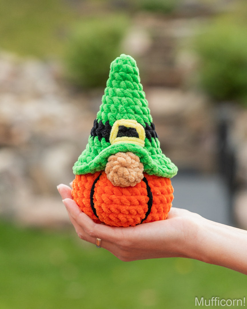 A crocheted gnome pumpkin held in a person's hand outdoors. The small decorative pumpkin has an orange body with black stitched lines, a tan textured nose, and a green pointed hat with black and yellow accents.