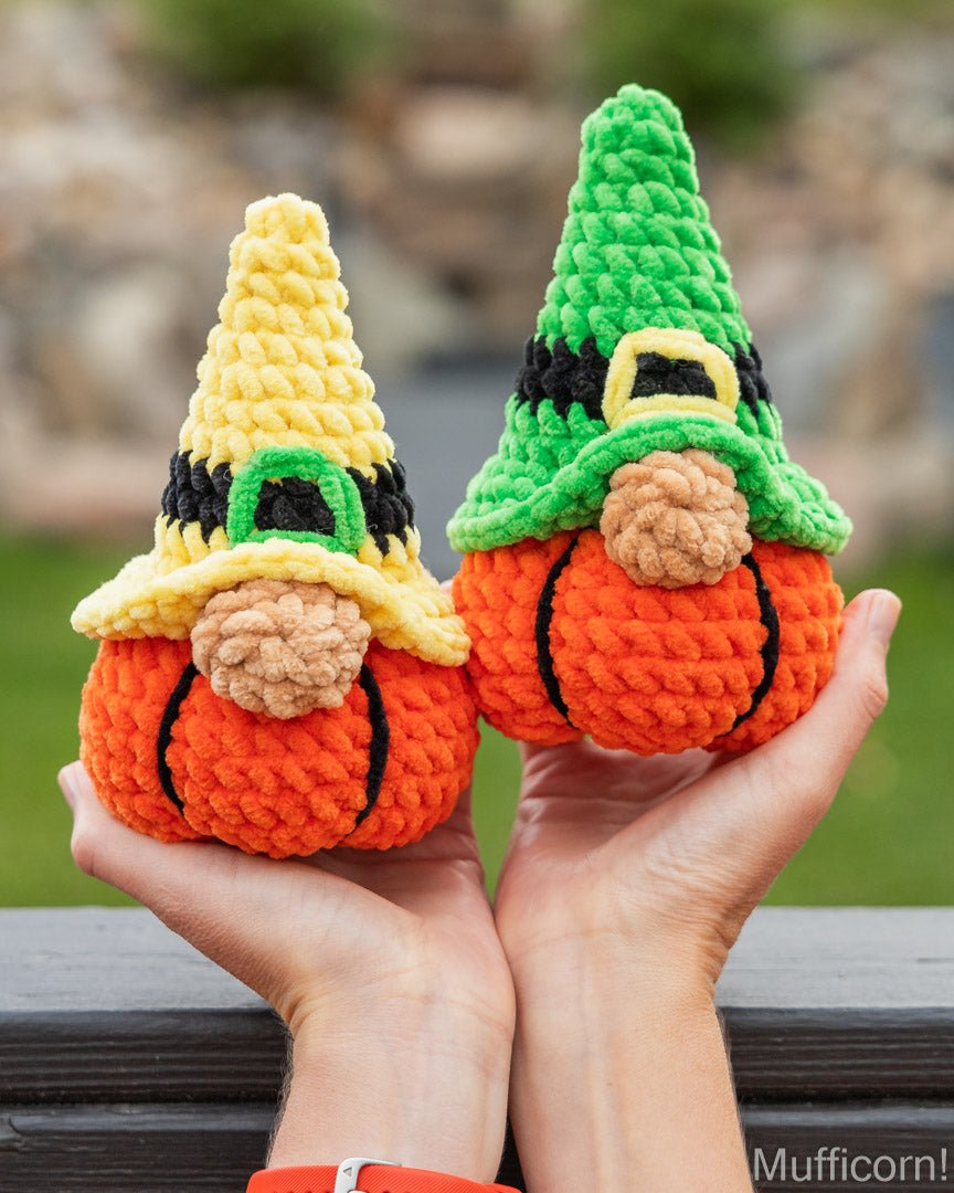 Two crocheted gnome pumpkins held in hands outdoors. One has a yellow hat with a green buckle and the other has a green hat with a yellow buckle.