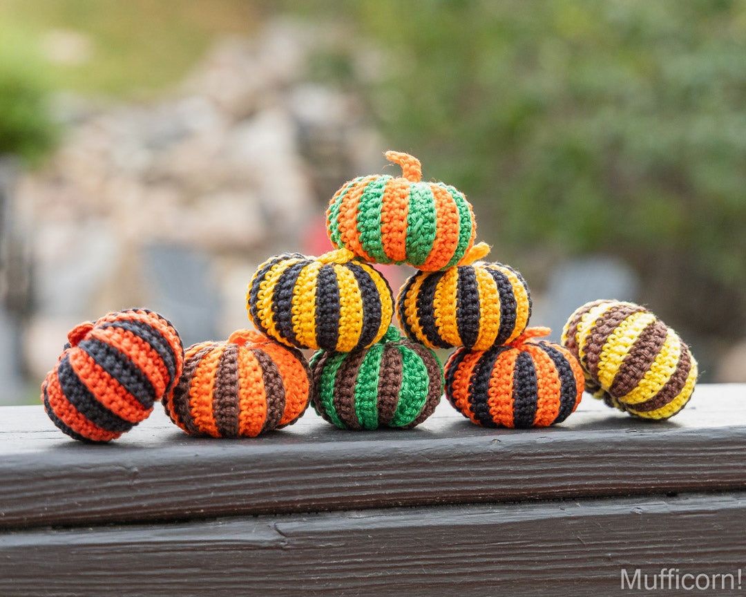 A group of crocheted decorative pumpkins in orange, green, yellow, and black stripes, arranged on a dark wooden surface with a blurred outdoor background.