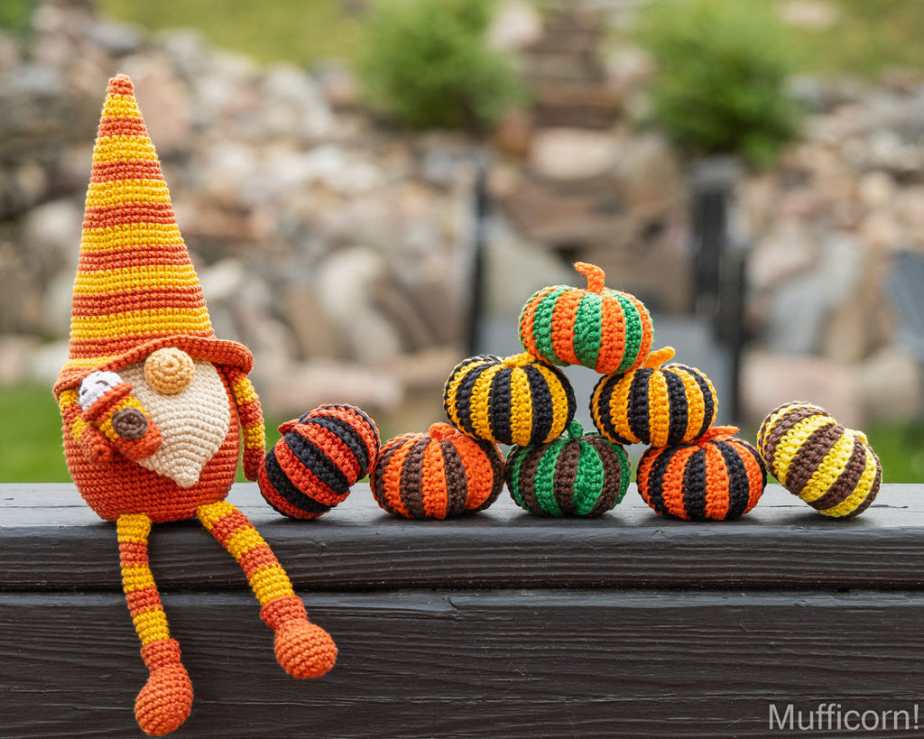 A crocheted gnome doll with a tall orange and yellow striped hat sits beside a small stack of crocheted pumpkins in orange, black, green, and yellow, placed on a dark wooden surface with a blurred outdoor garden background.