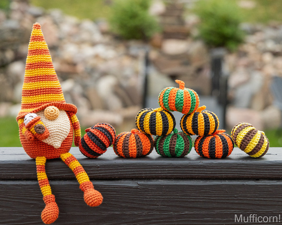 A crocheted gnome doll with a tall orange and yellow striped hat sits beside a small stack of crocheted pumpkins in orange, black, green, and yellow, placed on a dark wooden surface with a blurred outdoor garden background.